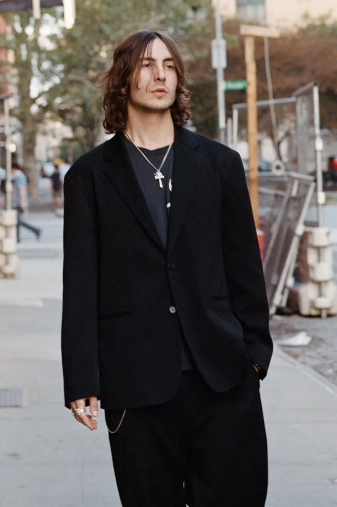 Long-haired person in a black suit and layered necklaces stands on a city sidewalk, one hand in a pocket