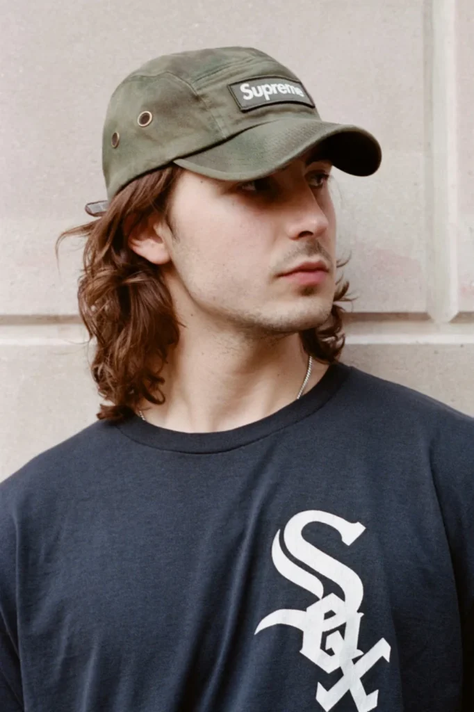 Person wearing an olive Supreme camp cap and navy T-shirt with a large white Sox logo, posed against a stone wall