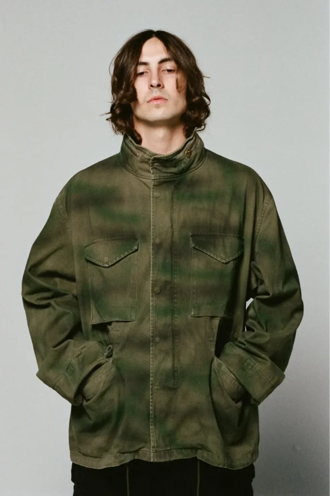 Person with long dark hair wears an olive field jacket with multiple flap pockets and hands in the pockets against a gray