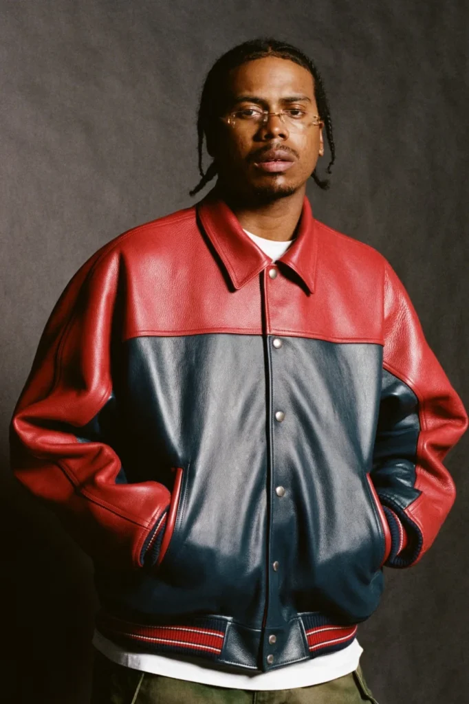 Man with braided hair and glasses wears a red and navy leather jacket, hands in pockets, against a dark backdrop