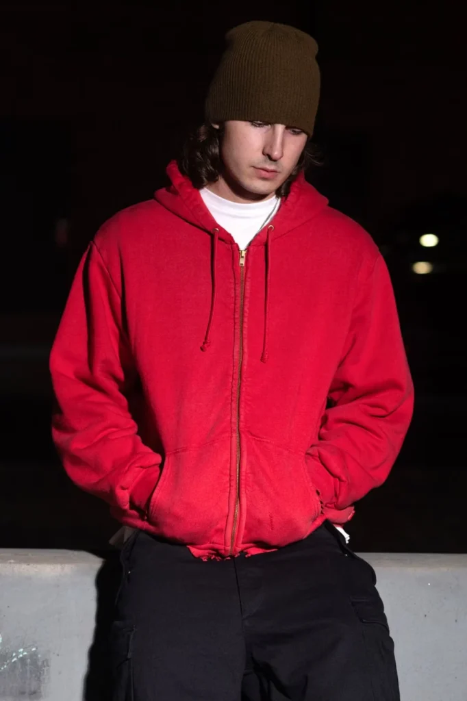 Model in a brown beanie and red zip-up hoodie stands against a dark background with hands in pockets