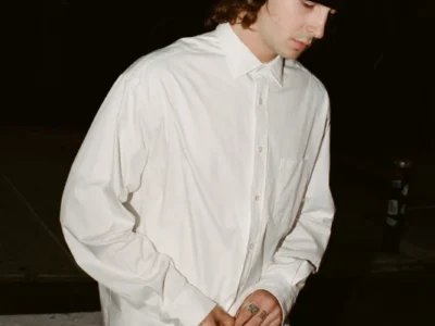Person in a black beanie buttons a white long-sleeve shirt at night outside a lit storefront