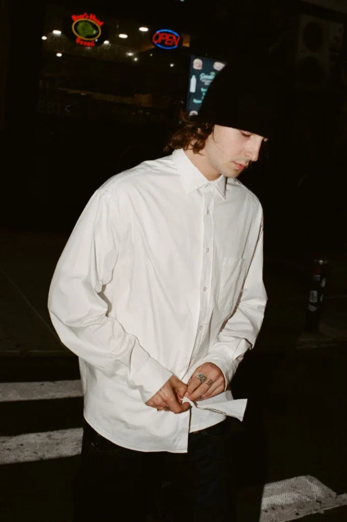 Person in a black beanie buttons a white long-sleeve shirt at night outside a lit storefront