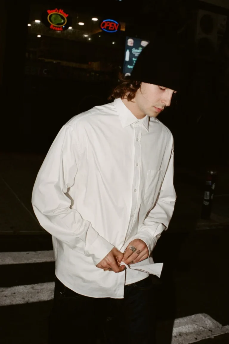 Person in a black beanie buttons a white long-sleeve shirt at night outside a lit storefront