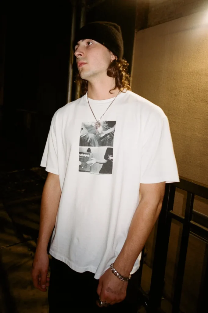 Person in a black beanie and white graphic T-shirt stands by a railing at night, lit against a building wall