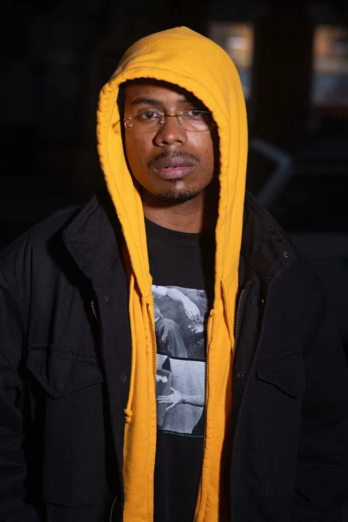 Man wearing glasses, a bright yellow hooded sweatshirt, black jacket, and graphic T-shirt against a dark background
