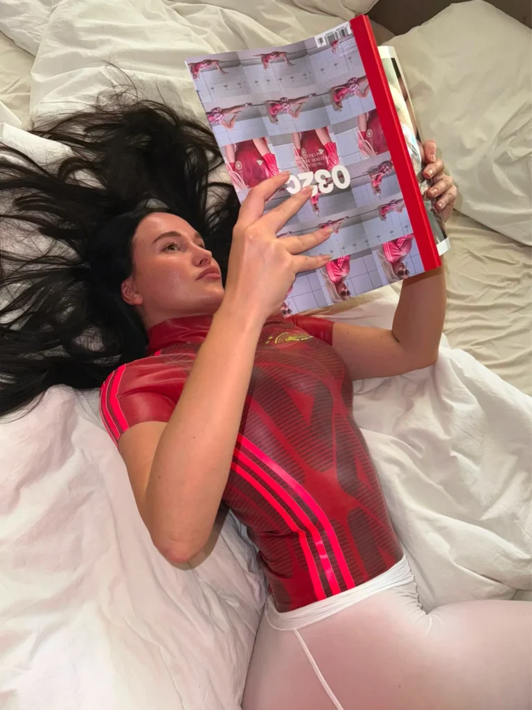 Jenna lies on white bedding in a red sheer striped top and white bottoms, holding a red fashion magazine over her face