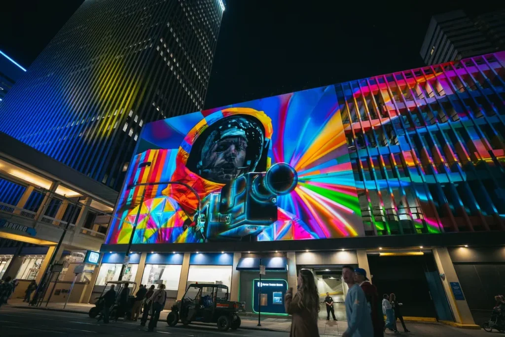 Colorful projection of an astronaut filming with a camera across a downtown building facade at night, with pedestrians below