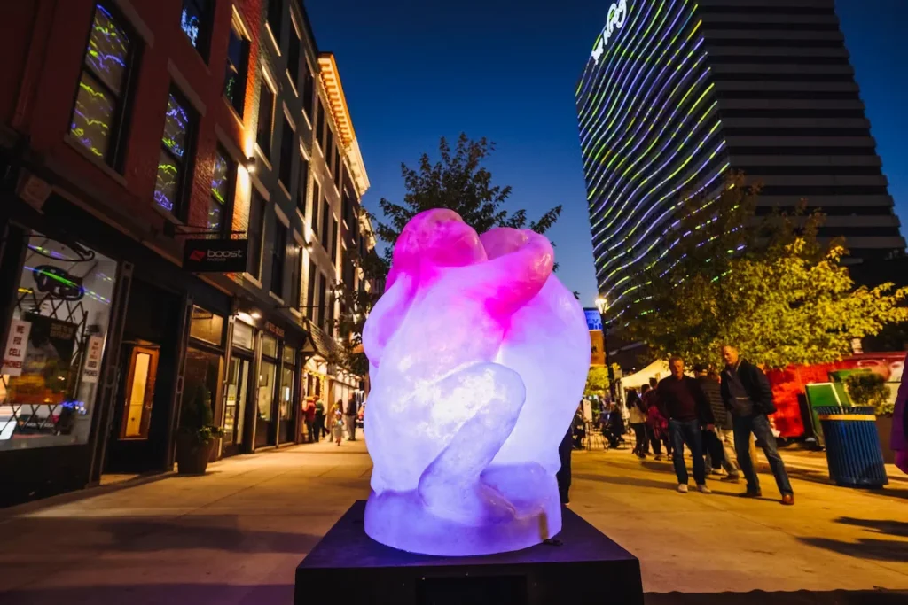 Glowing pink and lavender abstract sculpture on a pedestal along a downtown street, with illuminated buildings and