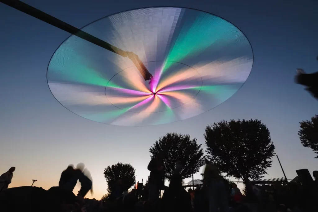 Large suspended oval canopy projects teal, pink and peach radial lights above silhouetted festivalgoers at dusk
