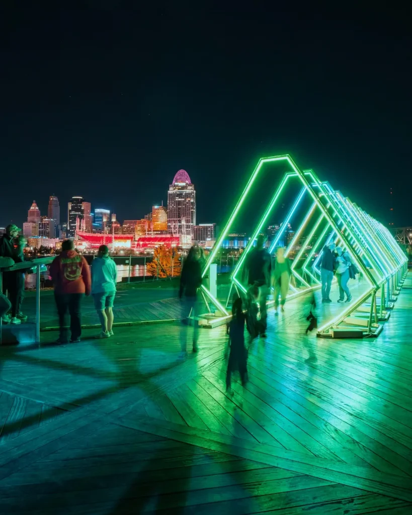 Visitors walk beneath glowing green and blue triangular light arches on a waterfront boardwalk, with Cincinnati’s skyline