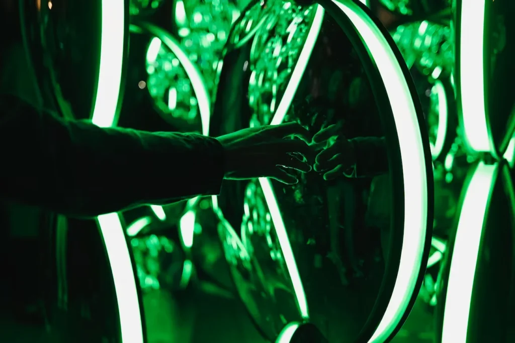Person reaches toward their reflection inside a dark installation of bright green glowing rings