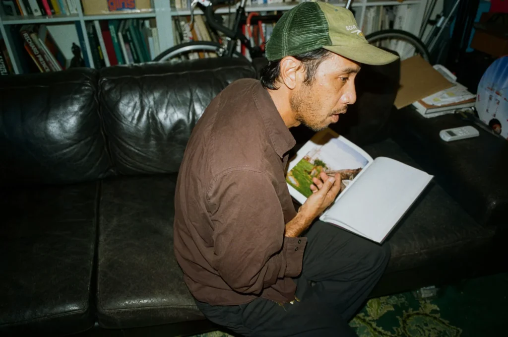 Jerry Hsu sits on a black leather couch, holding an open photo book, wearing a brown shirt and green cap
