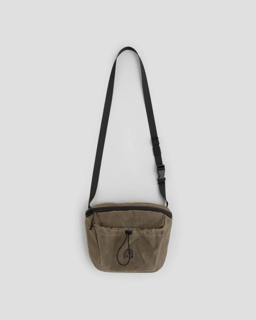 Brown canvas shoulder bag with black adjustable strap, zipper top, and front drawstring pocket on a light gray background