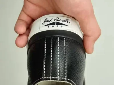 Close-up of a hand holding a black leather shoe, show white stitch and a cream heel tab mark Jack Purcell 1935