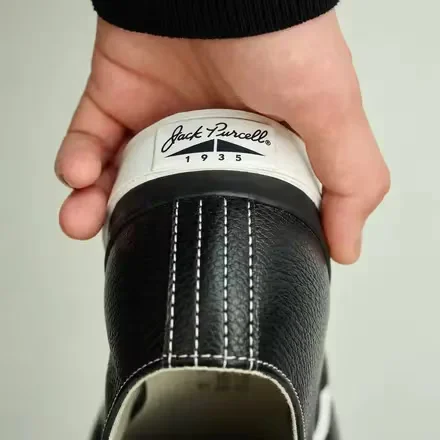 Close-up of a hand holding a black leather shoe, show white stitch and a cream heel tab mark Jack Purcell 1935
