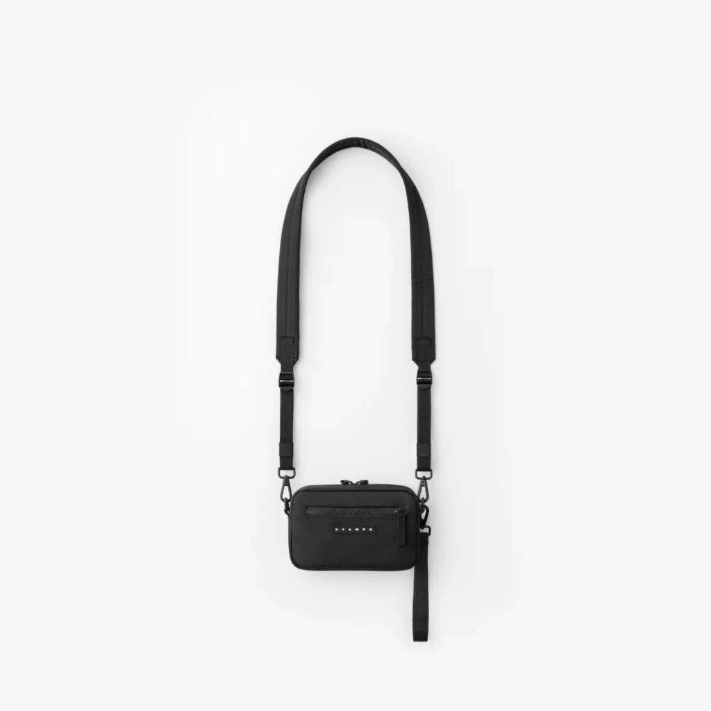 Black crossbody bag with adjustable strap, front zip pocket, logo patch, and side hanging strap on white background
