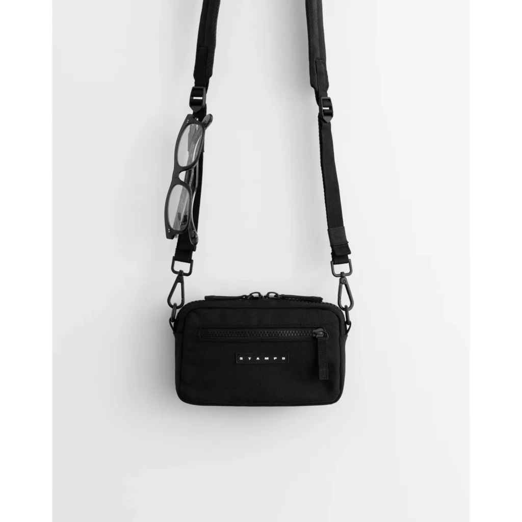 Black crossbody bag with adjustable strap, front zip pocket, and attached black eyeglasses, displayed against white