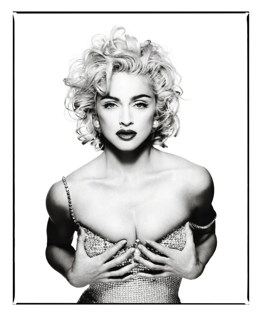 Black-and-white portrait of a woman with curly hair in a jeweled off-shoulder dress, hands across her chest, framed in white
