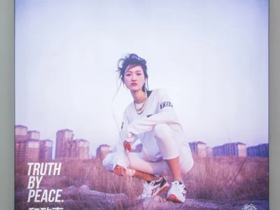 Person crouching in a dry field wearing white athletic clothes, sneakers and a gold chain, with apartment buildings behind