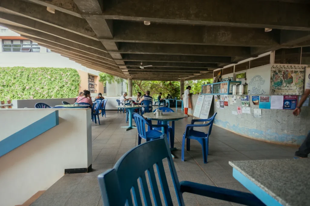 Open-air café with blue plastic chairs and tables beneath exposed concrete beams, diners seated beside a poster-covered