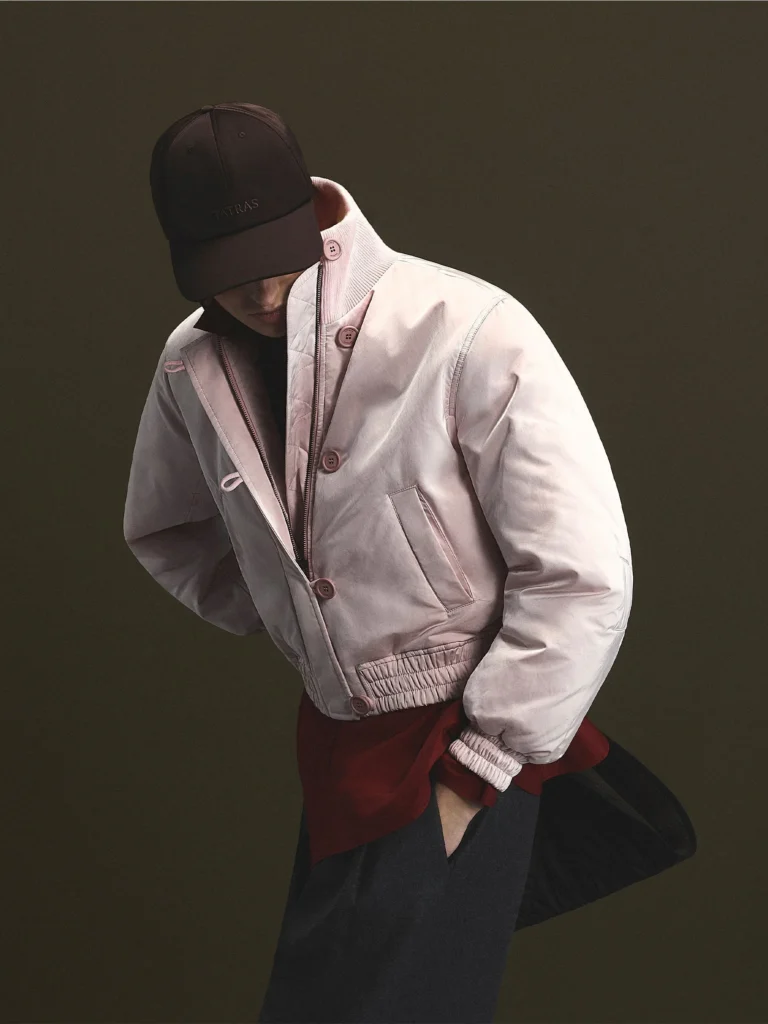 Person in a dark cap and pale pink quilt jacket lean forward against a dark olive background