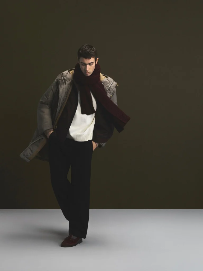 Model stands with one leg cross, wear a gray hooded jacket, burgundy scarf, white sweater, and black trousers against a
