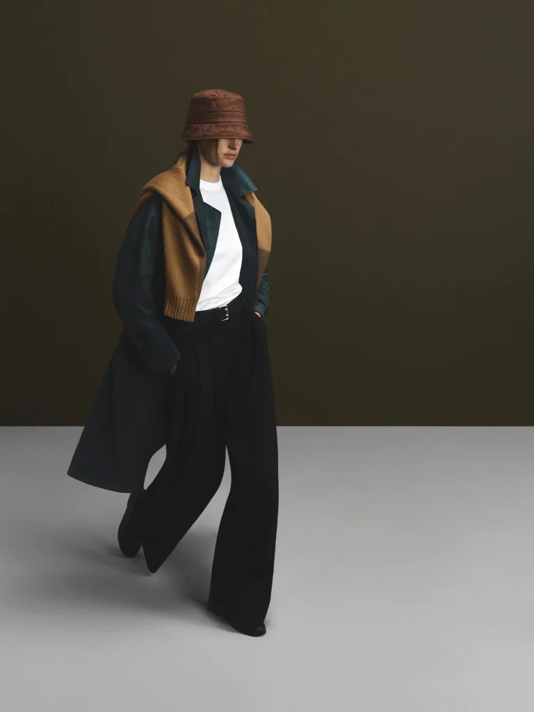 Model poses in a brown quilted bucket hat, brown knit vest, dark jacket, white shirt and wide black trousers against a dark