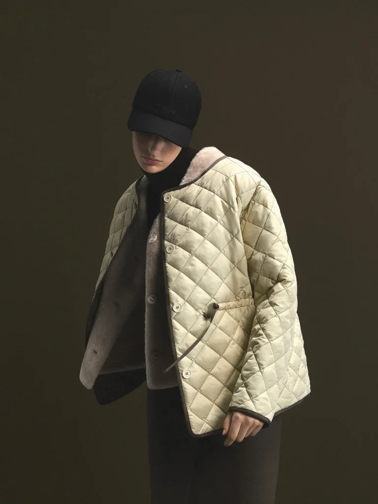 Model in a black cap and scarf wear a cream diamond-quilted jacket with shearling collar against a dark backdrop
