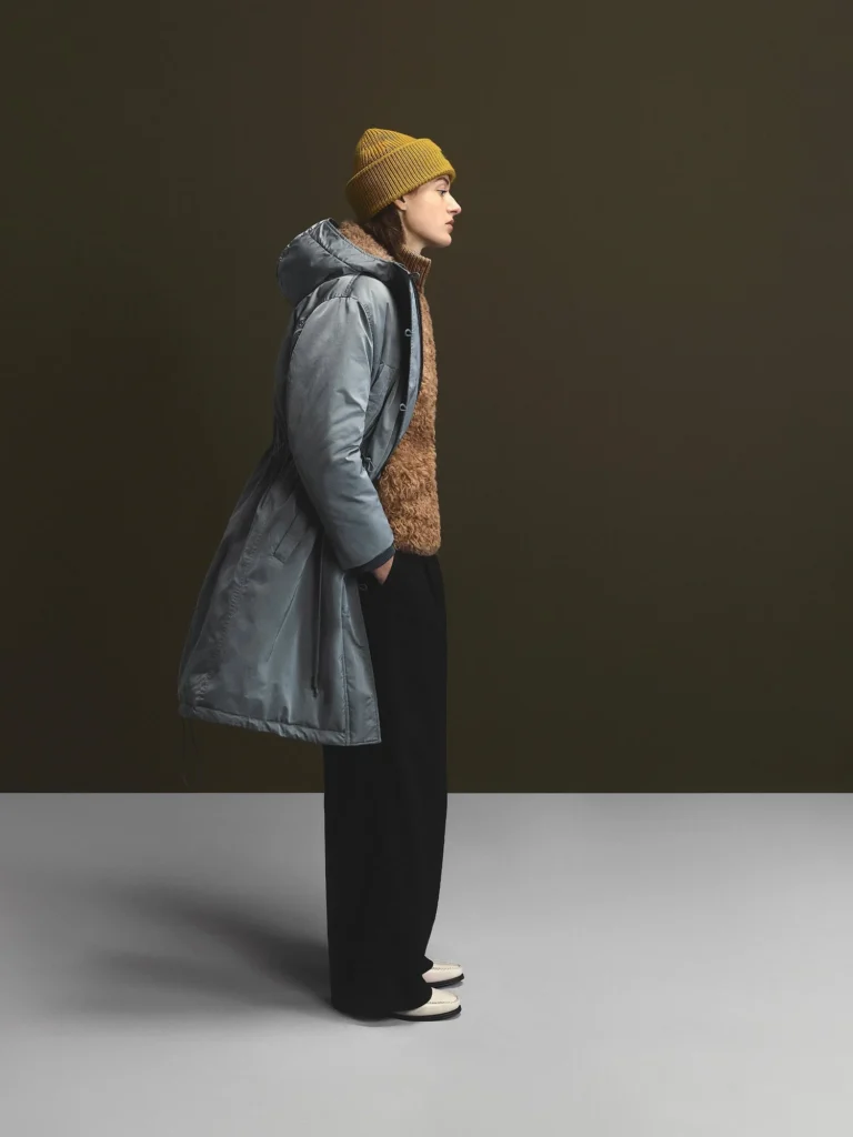 Side-profile model in a gray hooded parka, brown fuzzy vest and mustard beanie, standing against a dark studio wall
