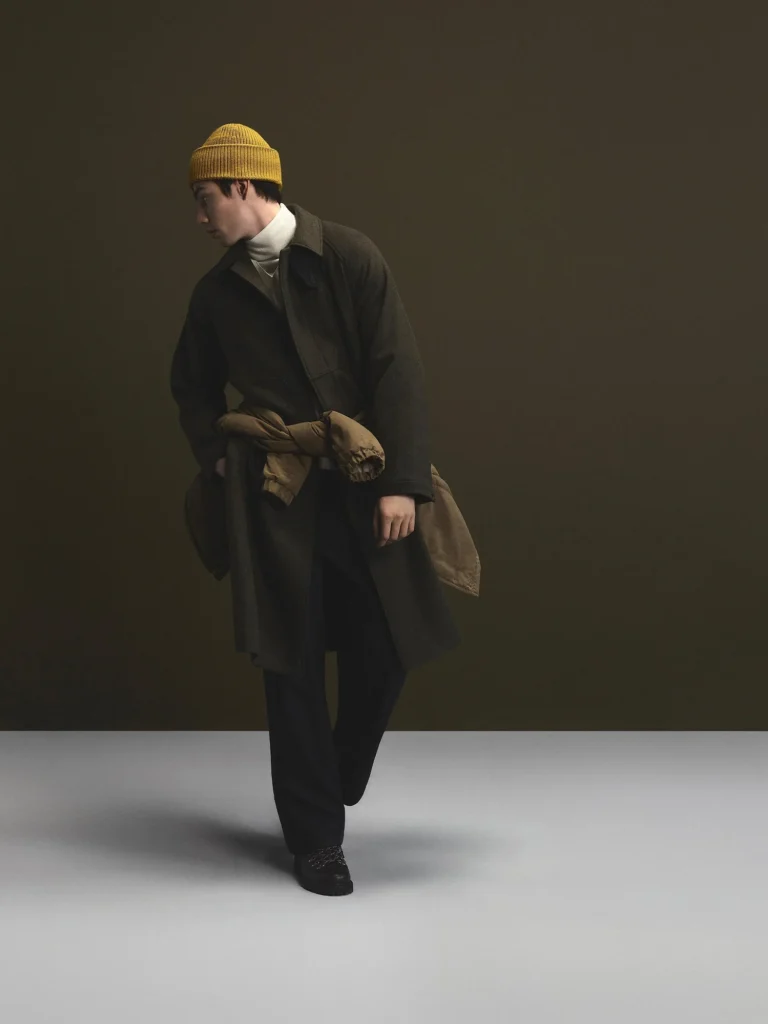 Model in a mustard beanie and dark coat poses against a dark backdrop, with a tan puffer jacket tied at the waist