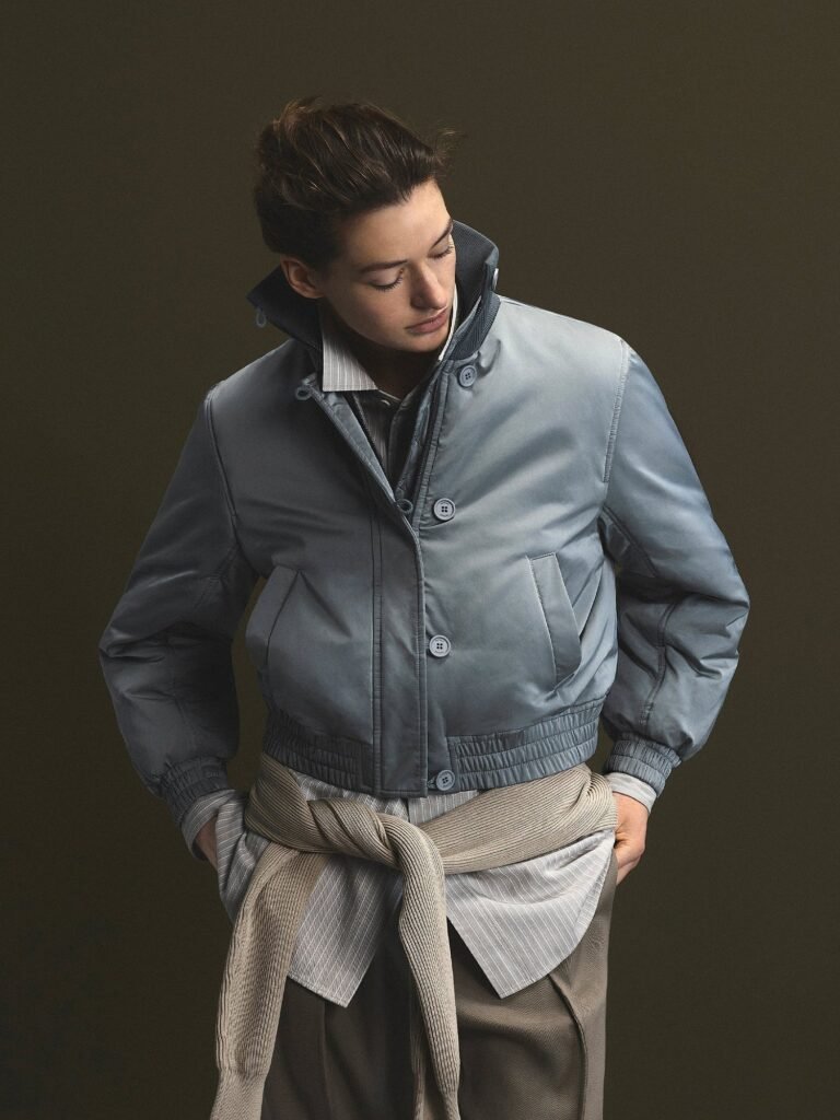 Model looks down wear a slate-gray cropped jacket with a high collar, layered shirts, and a beige sweater tied at the