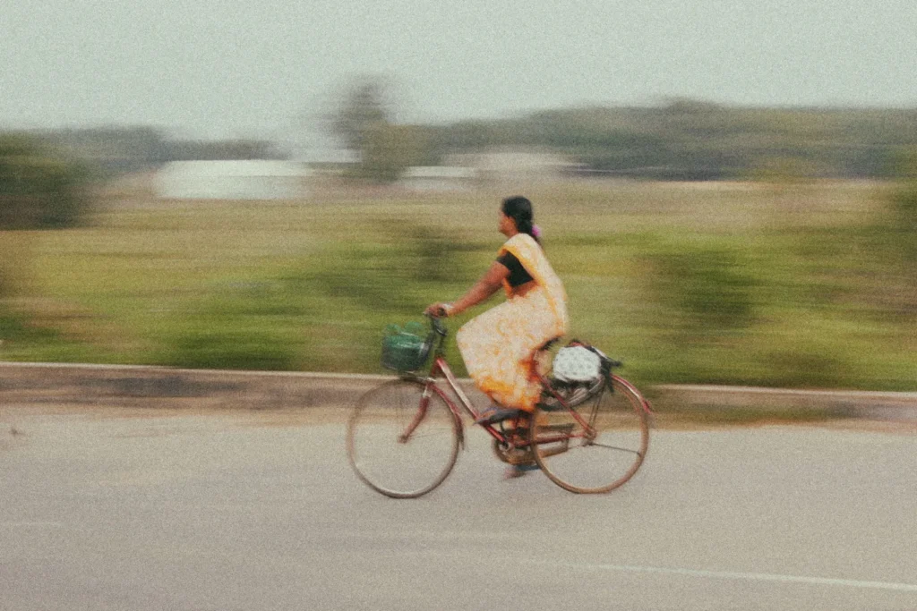 A woman in a yellow patterned sari rides a bicycle along a road beside a blurred grassy field and pond
