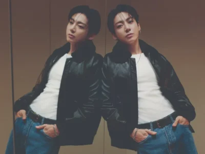 Jung Kook poses with hands in his jeans pockets beside his reflection, wearing a black leather jacket and white shirt in an