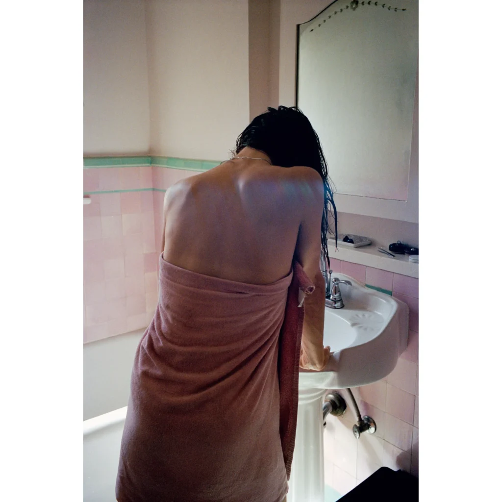 Person wrapped in a mauve towel leans over a white sink in a pink-and-mint tiled bathroom