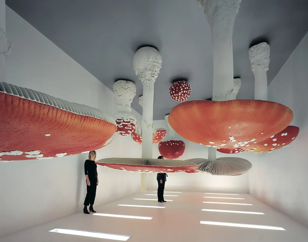 Visitors stand beneath giant red-and-white mushroom sculptures in a bright white gallery with illuminated floor panels