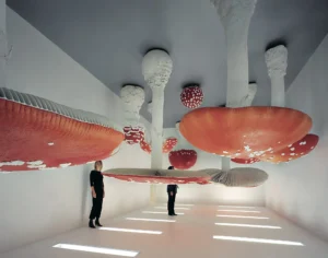 Visitors stand beneath giant red-and-white mushroom sculptures in a bright white gallery with illuminated floor panels