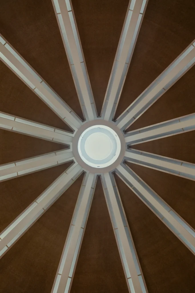 Upward view of a domed ceiling with white radial beams converging around a circular skylight against brown panels
