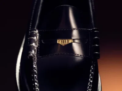 Close-up of a glossy black leather loafer with moccasin stitching and a gold 90s emblem on the vamp against a dark background