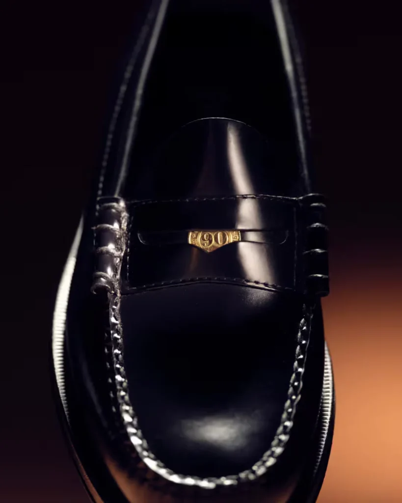Close-up of a glossy black leather loafer with moccasin stitching and a gold 90s emblem on the vamp against a dark background