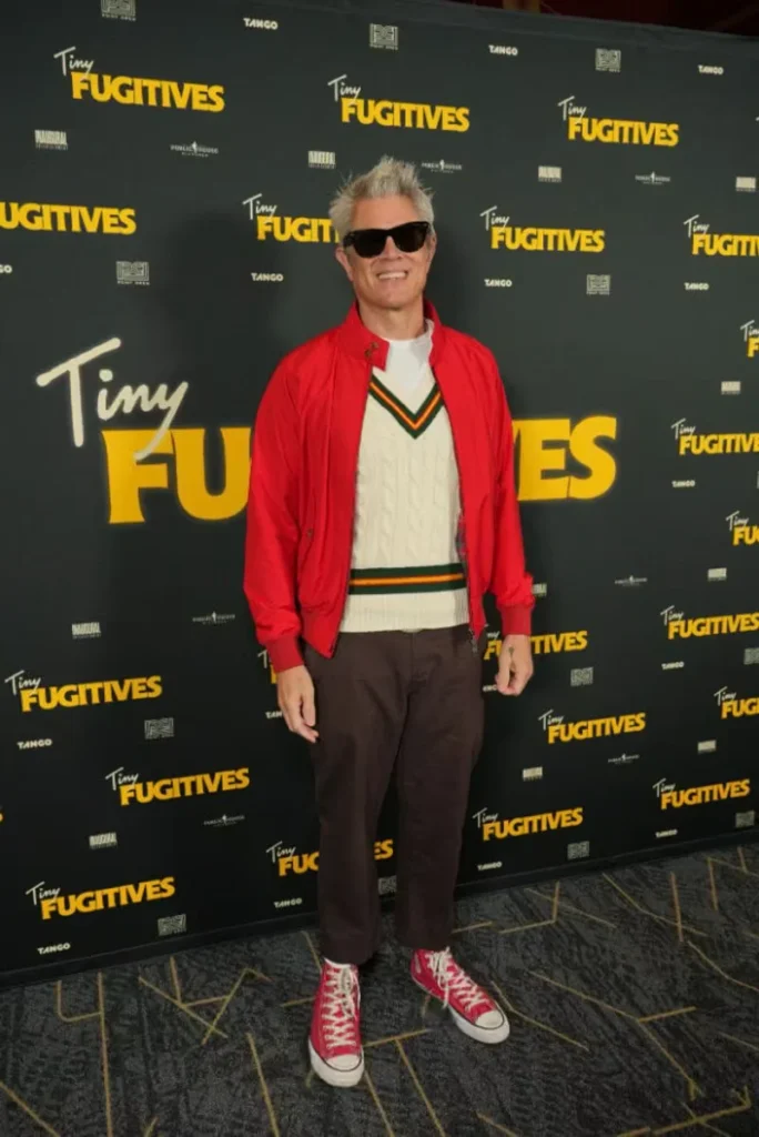 Johnny Knoxville poses in sunglasses, a red jacket, cream sweater vest, brown pants and red sneakers before a Tiny Fugitives