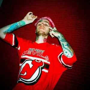 Tattooed Lil Peep with pink hair poses in a red New Jersey Devils hockey jersey against a dark red wall
