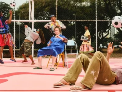 Kid Super Mid of group people by pool in tincture outfits play soccer on a pink court, with one person reclining in the foreground in star-patterned pants