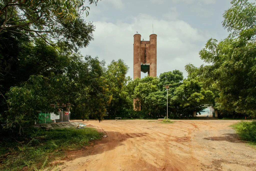 Red-brick tower with twin cylindrical columns rises above dense green trees beside an unpaved sandy road