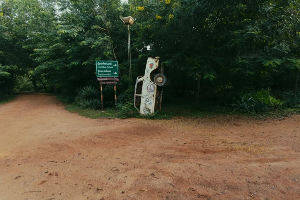 A weathered, graffiti-covered car stands upright beside a bilingual sign on a red dirt road amid dense greenery