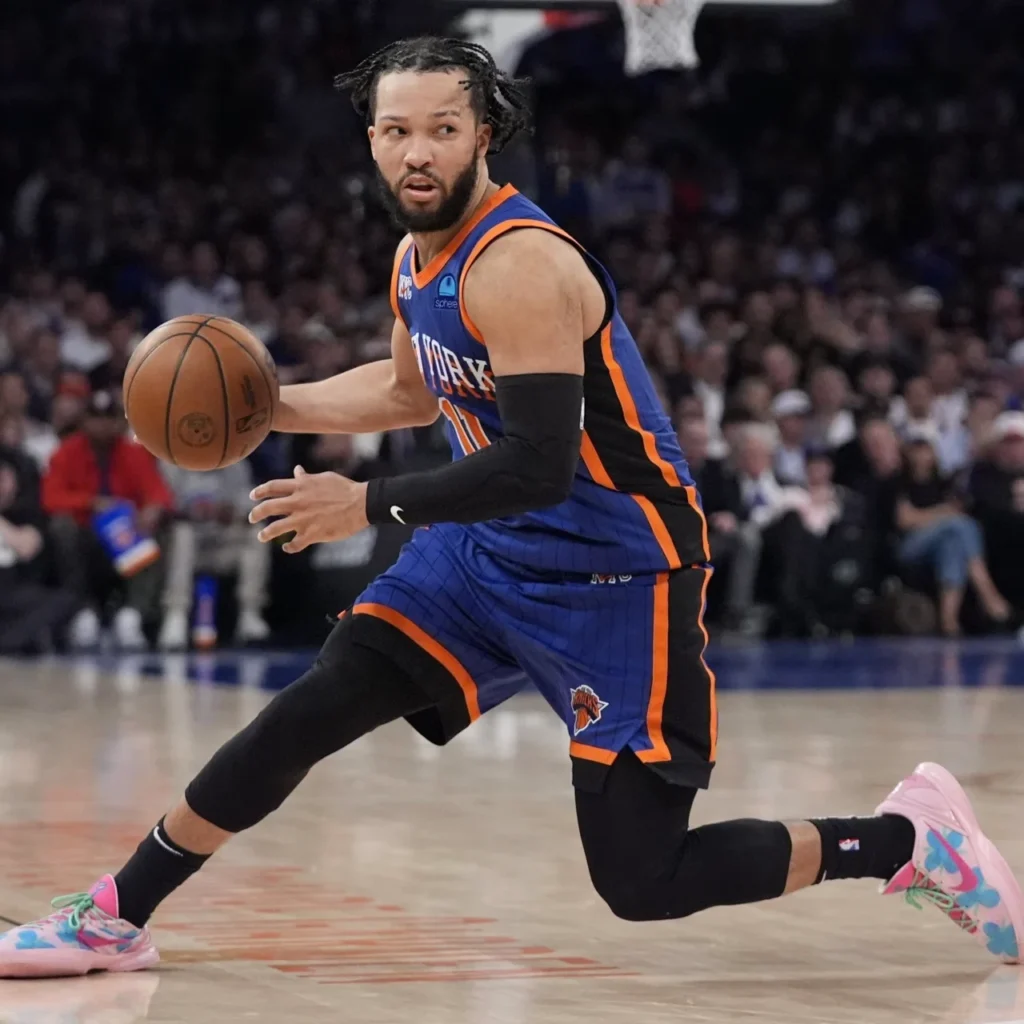Jalen Brunson dribbles a basketball while lunging across a court in a blue and orange Knicks uniform and pink sneakers