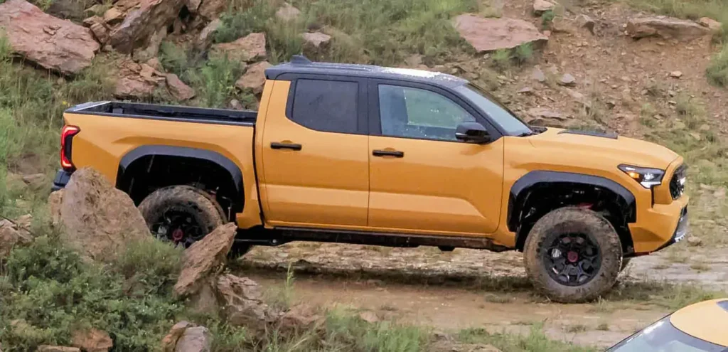 Orange pickup truck with black trim and muddy tires navigating rocky terrain beside a log