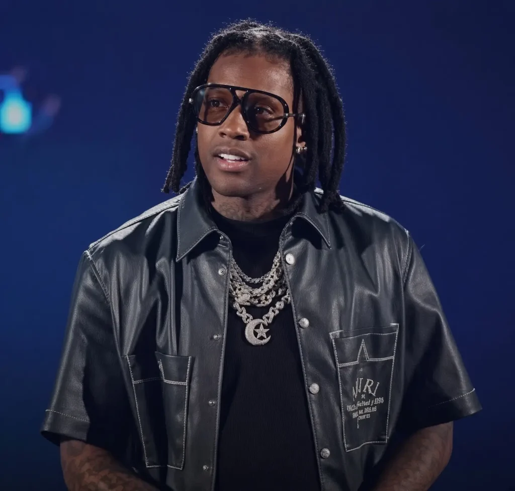Lil Durk wears oversized black glasses, a black leather shirt, layered diamond chains, and shoulder-length dreadlocks