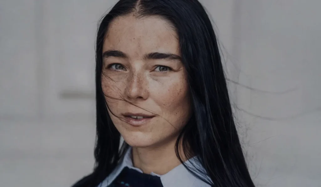 Close-up portrait of Julie Kegels with long dark hair, wearing a pale blue collared shirt against a gray background