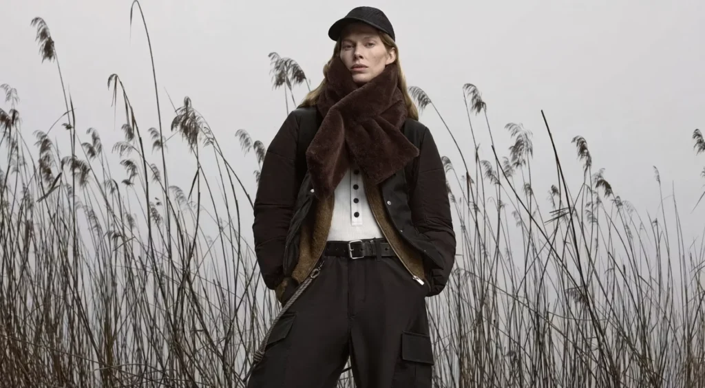 Model in black outerwear and cap stands among tall reeds, wearing a large brown scarf and belted cargo pants